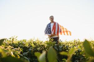 ein jung Farmer steht mit ein USA Flagge im ein Sojabohne Feld. das Konzept von das uns landwirtschaftlich Industrie. foto