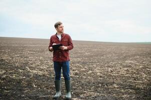 ein Farmer im Stiefel funktioniert mit seine Tablette im ein Feld gesät im Frühling. ein Agronom Spaziergänge das Erde, beurteilen ein gepflügt Feld im Herbst. Landwirtschaft. Clever Landwirtschaft Technologien foto