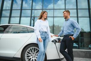 lächelnd Mann und Frau auf das Laden Bahnhof zum elektrisch Autos. ein Mann ist Laden ein Wagen. foto