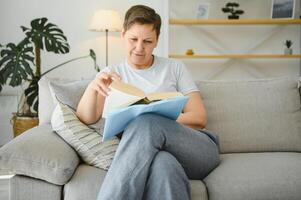 Porträt von ein glücklich reifen Frau entspannend auf Couch und lesen ein Buch im ein modern Haus foto