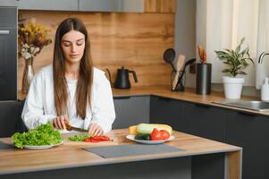 glücklich lächelnd süß Frau ist vorbereiten ein frisch gesund vegan Salat mit viele Gemüse im das Küche beim Zuhause und versuchen ein Neu Rezept foto