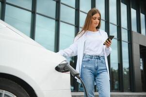 Telefon im Hände. Frau auf das elektrisch Autos aufladen Bahnhof beim Tageszeit. Marke Neu Fahrzeug. foto