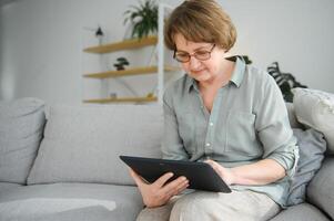 Pensionierung Freizeit. lächelnd Alten Frau ruhen mit Digital Tablette auf Couch beim heim, Surfen Internet oder Aufpassen Filme foto