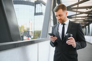 bärtig Geschäftsmann im formal passen auf brechen mit Handy, Mobiltelefon Telefon verwenden Smartphone. Geschäft Mann Stehen draußen auf modern städtisch Stadt Straße Hintergrund mit Kaffee Tasse im Innenstadt draußen. Kopieren Raum foto