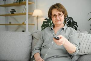 Alten Frau Herstellung Hören Zeichen, steigend Fernseher einstellen Volumen mit Fernbedienung Kontrolle. foto