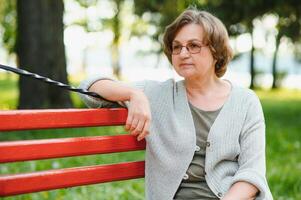 elegant Alten Frau im das Hemd ist Sitzung auf das Bank im ein Park auf ein warm Tag foto