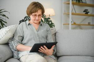 Pensionierung Freizeit. lächelnd Alten Frau ruhen mit Digital Tablette auf Couch beim heim, Surfen Internet oder Aufpassen Filme foto