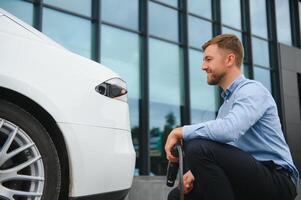 Porträt von jung gut aussehend Mann im beiläufig tragen, Stehen beim das Laden Bahnhof. Öko elektrisch Auto Konzept. foto