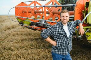 glücklich Farmer stolz Stehen im ein Feld. kombinieren Mähdrescher Treiber gehen zu Ernte Reich Weizen Ernte. Agronom tragen Flanell Shirt, suchen beim Kamera auf ein Ackerland foto