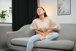 jung weiblich Leiden bilden Bauch schmerzen während Sitzung auf Couch beim heim. Ursachen von Bauch Schmerzen umfassen Menstruation Schmerz, Gastritis, Bauch Geschwür, Essen Vergiftung, Durchfall oder IBS. selektiv Fokus foto