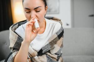 Nasal- sprühen im Hand von krank jung Frau Sitzung auf Couch. allergisch Rhinitis Symptome und Behandlung foto