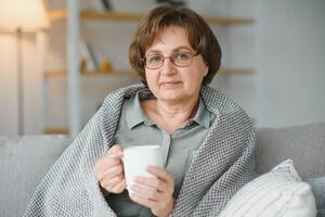lächelnd reifen ältere 65s Frau Sitzung entspannend mit Tasse von Tee, Kaffee. Senior Mitte Alter stilvoll aussehen Frau mit Brille Porträt mit Tasse suchen Weg beim modern Zuhause foto