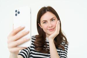 Porträt von jung europäisch Frau nehmen Selfie auf Smartphone, halten Weiß Handy, Mobiltelefon Telefon und posieren zum Fotos, isoliert gegen Weiß Hintergrund foto