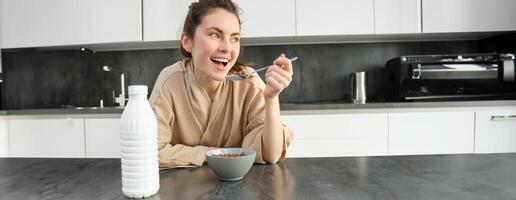 Porträt von jung schön Frau im Bademantel, Essen Getreide zum Frühstück, lehnt sich auf Küche Arbeitsplatte, suchen beim ihr Morgen Mahlzeit foto
