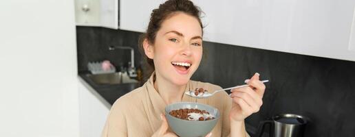 glücklich Morgens. Herrlich jung Frau Essen Getreide mit Milch, Stehen im Küche mit Frühstück Schüssel, genießen Start von das Tag, lächelnd foto