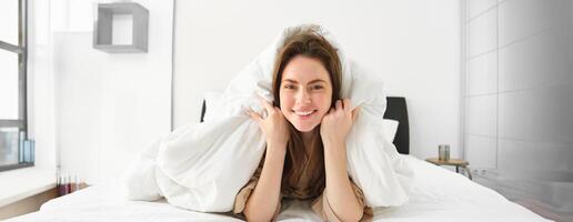 süß Mädchen mit unordentlich Haar, Lügen im Bett bedeckt im Weiß Blätter Bettdecke, lächelnd und Lachen kokett, Ausgaben Zeit im ihr Schlafzimmer foto