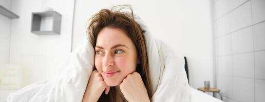 süß Mädchen mit unordentlich Haar, Lügen im Bett bedeckt im Weiß Blätter Bettdecke, lächelnd und Lachen kokett, Ausgaben Zeit im ihr Schlafzimmer foto