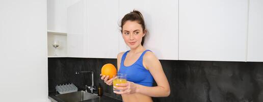 Innen- Schuss von jung Brünette Frau im Sportbekleidung, Trinken Orange Saft, halten Obst im Hand, posieren im Küche foto