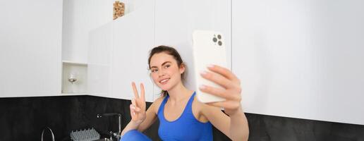 Porträt von schön Frau, Fitness Blogger, nehmen Selfie im sportbra und Gamaschen, posieren im Küche mit Smartphone foto