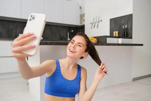 schließen oben Porträt von jung sportlich Frau, Fitness Mädchen nimmt Selfie beim Zuhause während tun ihr trainieren, posieren auf Yoga Matte, hält Smartphone und lächelt foto