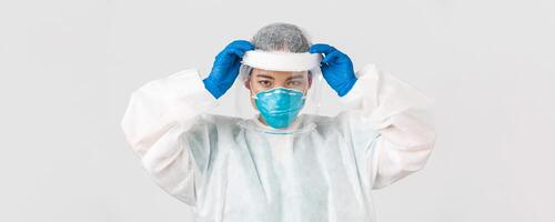 COVID-19, Coronavirus Krankheit, Gesundheitswesen Arbeitskräfte Konzept. zuversichtlich ernst aussehend asiatisch weiblich Arzt, stellen auf Gesicht Schild und persönlich schützend Ausrüstung Vor eintreten ansteckend Zone foto