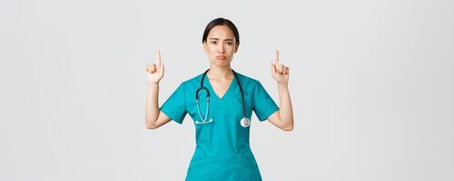 COVID-19, Gesundheitswesen Arbeitskräfte, Pandemie Konzept. düster enttäuscht asiatisch weiblich Arzt, Arzt im Peelings zeigen Finger oben und verzog das Gesicht unzufrieden, Stehen Weiß Hintergrund foto