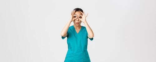 COVID-19, Gesundheitswesen Arbeitskräfte und verhindern Virus Konzept. beeindruckt komisch und süß weiblich asiatisch Krankenschwester im Peelings suchen durch Finger Über Augen wie wenn starren im Ferngläser erstaunt foto