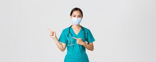 COVID-19, Coronavirus Krankheit, Gesundheitswesen Arbeitskräfte Konzept. Fachmann zuversichtlich asiatisch weiblich Arzt, Arzt im medizinisch Maske und Peelings zeigen Finger links, zeigen Werbung foto