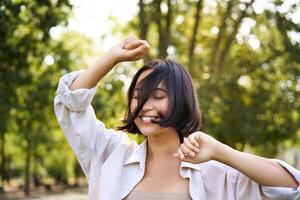 Menschen Lebensstil. Porträt von jung Brünette Frau Tanzen, lächelnd und Lachen, Gehen im Park mit Hände Aufzug oben hoch, genießen Sommer- Tag draußen foto