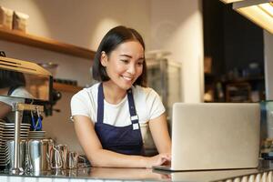 Porträt von lächelnd asiatisch Barista, Cafe Inhaber Unternehmer, Arbeiten auf Laptop, wird bearbeitet Aufträge auf Computer, Stehen hinter Zähler foto