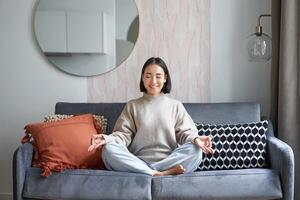 Entspannung und Geduld. lächelnd jung asiatisch Frau im gemütlich Zimmer, Sitzung auf Sofa und meditieren, tun Yoga Achtsamkeit Ausbildung foto