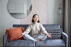 Entspannung und Geduld. lächelnd jung asiatisch Frau im gemütlich Zimmer, Sitzung auf Sofa und meditieren, tun Yoga Achtsamkeit Ausbildung foto