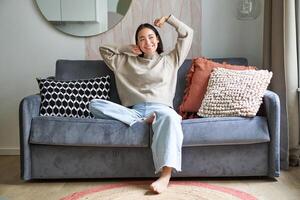 Porträt von glücklich asiatisch Frau Gefühl faul, Dehnen auf Sofa und lächelnd erfreut, entspannend beim heim, ruhen von Arbeit foto