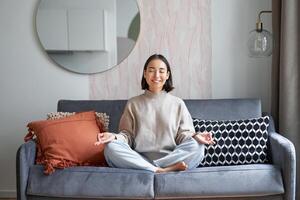Entspannung und Geduld. lächelnd jung asiatisch Frau im gemütlich Zimmer, Sitzung auf Sofa und meditieren, tun Yoga Achtsamkeit Ausbildung foto