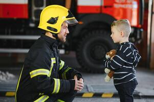 Porträt von gerettet wenig Junge mit Feuerwehrmann Mann Stehen in der Nähe von Feuer LKW. Feuerwehrmann im Feuer Kampf Betrieb. foto