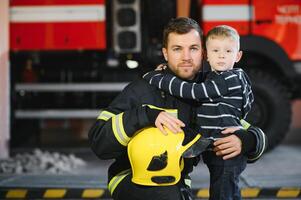 Porträt von gerettet wenig Junge mit Feuerwehrmann Mann Stehen in der Nähe von Feuer LKW. Feuerwehrmann im Feuer Kampf Betrieb. foto