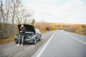 Mann reparieren ein gebrochen Auto durch das Straße. Mann haben Ärger mit seine gebrochen Auto auf das Autobahn Straßenrand. Mann suchen unter das Auto Haube. Auto geht kaputt Nieder auf das Autobahn. Straßenrand Hilfe Konzept. foto