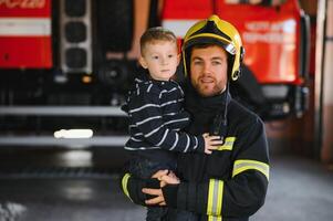 ein Feuerwehrmann nehmen ein wenig Kind Junge zu speichern ihn. Feuer Motor Auto auf Hintergrund. Feuerwehrmann mit Kind im seine Waffen. Schutz Konzept. foto