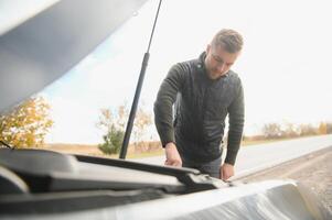 Mann reparieren ein gebrochen Auto durch das Straße. Mann haben Ärger mit seine gebrochen Auto auf das Autobahn Straßenrand. Mann suchen unter das Auto Haube. Auto geht kaputt Nieder auf das Autobahn. Straßenrand Hilfe Konzept. foto