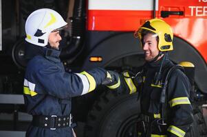 zwei Feuerwehrleute im schützend Kleidung im Helme mit Feuer Motor, freundlich Handschlag foto