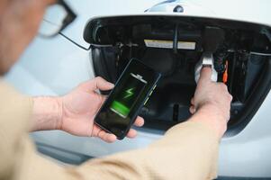 Mann Laden elektrisch Fahrzeug mit Kabel suchen beim App auf Handy, Mobiltelefon Telefon foto