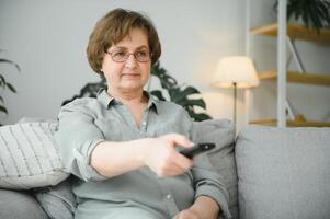 brechen Nachricht. schockiert Senior Frau Aufpassen Fernseher beim heim, Sitzung auf Couch mit Fernbedienung Regler im Hände foto