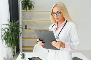 weiblich Arzt tragen Labor Mantel und Stethoskop und halten Zwischenablage im ihr Hände während Stehen beim Krankenhaus. foto