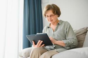 Pensionierung Freizeit. lächelnd Alten Frau ruhen mit Digital Tablette auf Couch beim heim, Surfen Internet oder Aufpassen Filme foto