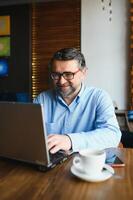 positiv Senior bärtig Mann mit grau Haar Trinken Kaffee und mit Laptop beim Cafe, Kopieren Raum. stilvoll alt Geschäftsmann im Burgund Jacke genießen seine Tee während Arbeiten online. foto