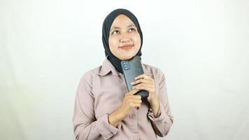 Porträt verwirrt jung asiatisch Frau im Hijab lächelnd Denken etwas während halten Handy, Mobiltelefon Telefon foto