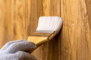 Hand im Handschuh halten Bürste ist bewirbt sich Lack oder schützend Lack auf hölzern Mauer. schützen das Holz von Feuchtigkeit. foto