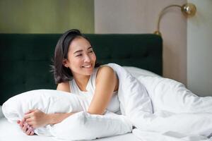 glücklich Morgens. schön Koreanisch Frau Lügen im Bett, lächelnd, aufwachen oben optimistisch, Startseite Sie selber mit warm Bettdecke foto