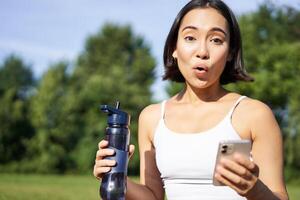 aufgeregt jung asiatisch Frau trainieren und Getränke Wasser, folgt Sport App auf Smartphone, sieht aus erstaunt und Glücklich, tut Übungen im Park foto