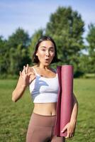 aufgeregt jung Frau Stehen mit Sport Matte, Yoga Kleidung, zeigt an okay Zeichen, trainieren im Park, Wohlbefinden Ausbildung Session draußen foto